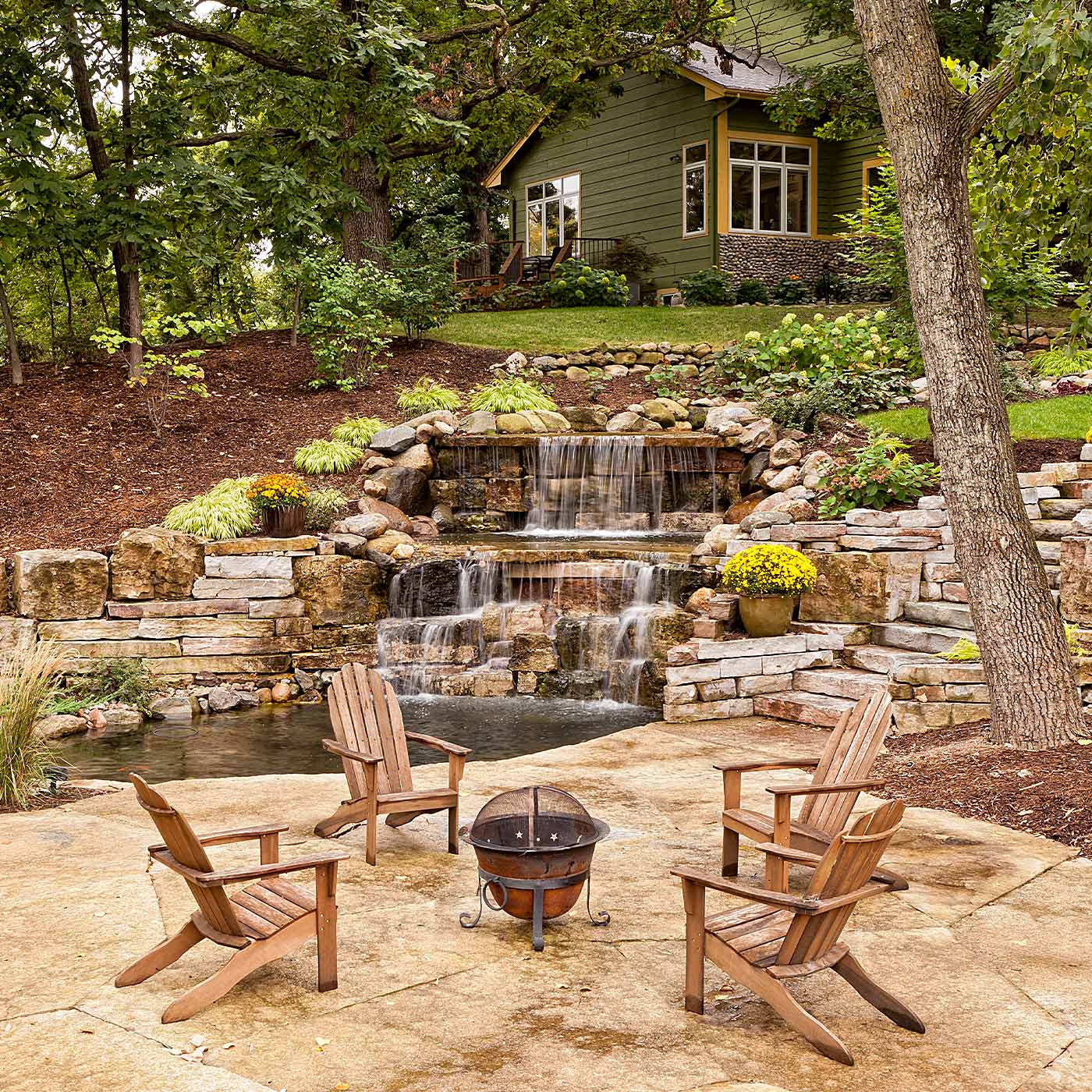 wooded chairs and a fire pit sit in front on a landscaped water fall in front of a green house up on the hill in Kensington, MD